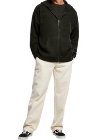 Svart ribbad hoodie med framdragkedja, texturerat tyg, kängurufickor, matchad med ljusbeige byxor och svarta sneakers.