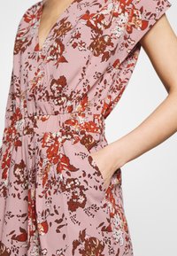 Vaaleanpunainen kukkakuosiset haalari, jossa on lyhyet hihat, V-mallinen kaula-aukko, joustava vyötärö ja sivutaskut. Sisältää punaisia ja ruskeita kukkakuvioita vaalealla taustalla.