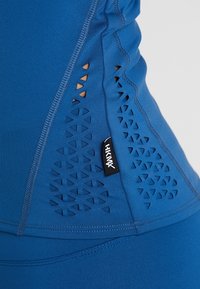 Modrý sportovní top bez rukávů s výřezovými vzory po stranách, hladkou texturou a malou značkovou nášivkou. Bezešvé provedení.