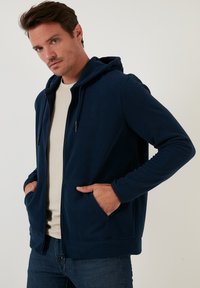 Felpa con cappuccio in pile blu navy, con zip frontale, cappuccio regolabile con cordino e due tasche laterali, che presenta un discreto logo sul petto.