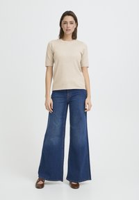 Pull beige à manches courtes associé à un jean en denim foncé à jambes larges. Des chaussures plates marron complètent la tenue, avec un fond uni.