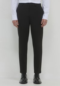 Uomo in pantaloni neri slim-fit, camicia bianca e scarpe da dress nere lucide in piedi contro uno sfondo bianco.