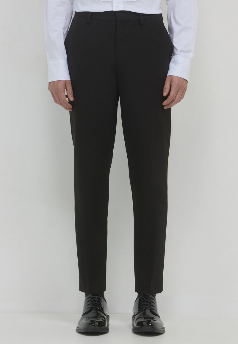 Uomo in pantaloni neri slim-fit, camicia bianca e scarpe da dress nere lucide in piedi contro uno sfondo bianco.