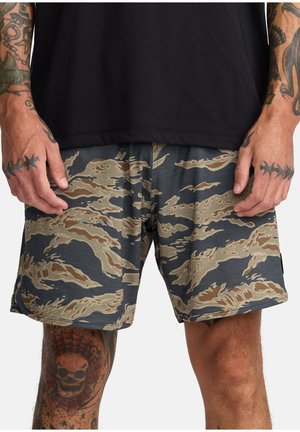 Man draagt camouflagepatroon korte broek en een zwart shirt, toont getatoeëerde handen en benen met schedel- en prikkeldraadontwerpen.