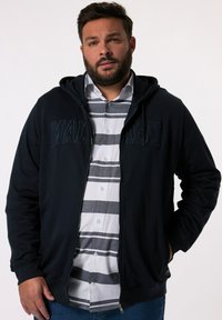 Marinblå zip-up hoodie med tryck framtill, dragsko i huvan, ribbade ärmslut, och en grå, vit och marinblå randig skjorta under.