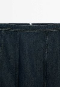 Jeanskjol i en mörkblå nyans, med veck i midjan och kontrasterande gula sömmar längs med sömmarna. Dragkedja bak.