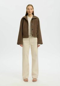 Femme debout portant une veste marron boutonnée, une chemise beige à col, un pantalon ample crème et des sandales beiges à bout ouvert sur un fond blanc.