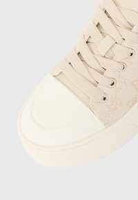 Μπεζ αθλητικά παπούτσια με υφασμάτινους κορδόνια, μεταλλικές θηλιές, καπάκι από καουτσούκ με υφή και διακριτικό μοτίβο στο πλάι, σε λευκό φόντο.