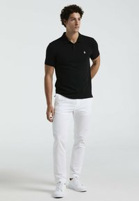 Zwarte poloshirt met een kraag, gedragen met witte slim-fit broek en witte sneakers. Heeft een klein logo op de linkerborst.