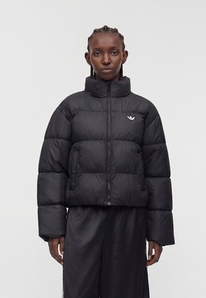 Persona con capelli intrecciati che indossa una giacca imbottita nera con cerniera e logo Adidas, abbinata a pantaloni neri larghi, in piedi davanti a uno sfondo semplice.