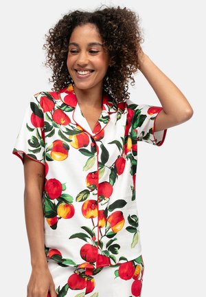 Donna sorridente con capelli ricci che indossa una camicia bianca a maniche corte con bottoni e pantaloni abbinati con stampa di mele rosse e gialle e foglie verdi.