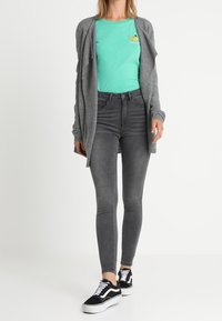 Grå stickad kavaj över en mintgröna t-shirt med en banangrafik, tillsammans med grå smala jeans och svarta sneakers med vita sulor.