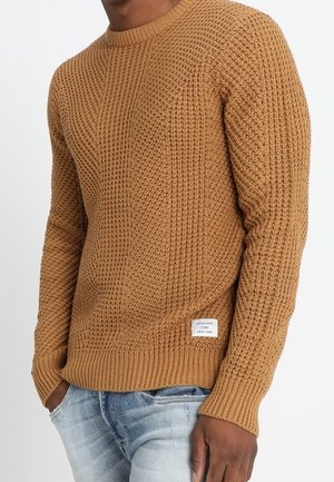 Homme portant un pull en tricot marron texturé et un jean bleu clair, les mains partiellement dans les poches, sur un fond blanc uni.