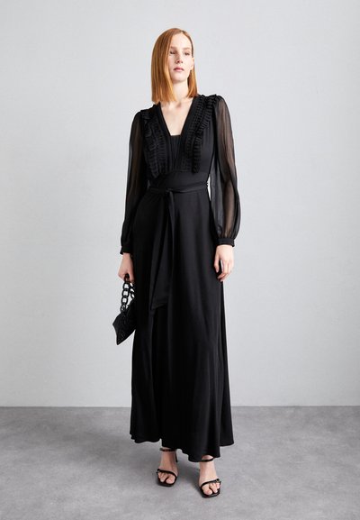 Robe noire à manches longues avec un col à volants, une jupe fluide et une ceinture à nouer. Associée à des sandales à lanières et un petit sac texturé.