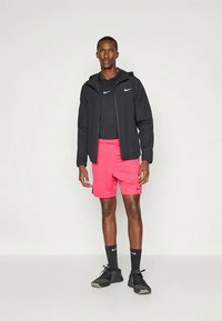 Musta vetoketjullinen urheilutakki, musta lyhythihainen paita, kirkkaanpinkit shortsit, mustat sukat, mustat urheilukengät. Nike-logo vaatteissa.