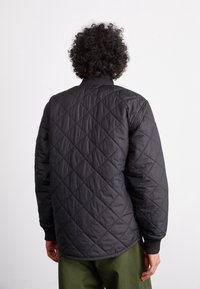 Sort quiltet jakke med strikket krave og manchetter, der har et struktureret diamantmønster. Bagfra ses en afslappet pasform og rene linjer.