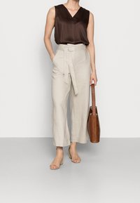 Haut marron sans manches, pantalon en lin beige clair taille haute avec ceinture à nouer, talons transparents, et sac fourre-tout en cuir marron.