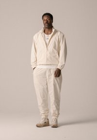 Veste zippée beige clair et joggers assortis en tissu texturé, portés avec un t-shirt blanc en dessous et des sneakers crème. Accents en chaîne dorée.