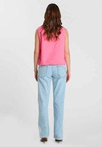 Femme aux cheveux bruns ondulés portant un haut rose sans manches, un jean bleu clair droit et des chaussures à talons marron, debout de dos.