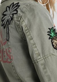 Oliivinvihreä takki, jossa on brodeerattu palmu ja kimalteleva ananas -pätkä. Kangas on sileä, näkyvillä saumoilla ja rento leikkaus.