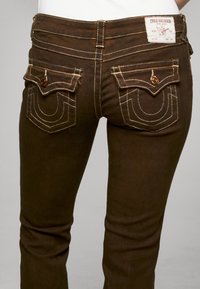 Brązowe dżinsy z denimu z tylnymi kieszeniami ozdobionymi charakterystycznymi wzorami przeszyć oraz z markową etykietą na pasku. Widoczna jest również faktura tkaniny.