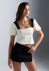Top court en crème avec des accents de nœud noir sur le décolleté carré, associé à une mini-jupe noire. Tissu lisse, design ajusté.