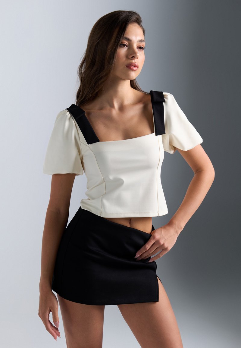 Top court en crème avec des accents de nœud noir sur le décolleté carré, associé à une mini-jupe noire. Tissu lisse, design ajusté.