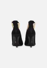 Tacchi alti in suede neri con punta affusolata, sottile tacco a spillo e dettagli in catena dorata sul retro per un tocco extra.