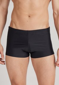 Schwarze Badehosen aus glattem, dehnbarem Material, mit einem niedrigen Schnitt und einem kleinen Logoetikett an der Seite.