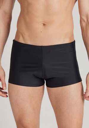 Schwarze Badehosen aus glattem, dehnbarem Material, mit einem niedrigen Schnitt und einem kleinen Logoetikett an der Seite.