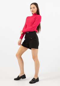 Červený žebrovaný rolák v kombinaci s černými šortkami s vysokým pasem, které mají přední kapsy. Model má na sobě černé mokasíny. Bílý pozadí.