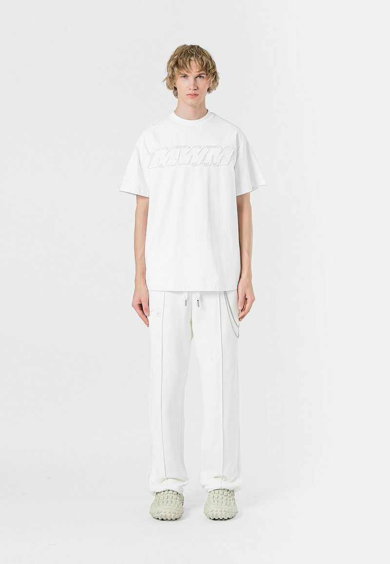 Camiseta blanca de manga corta con logo en relieve, combinada con pantalones blancos con bolsillos laterales y detalle en cadena, llevada con zapatos texturizados.