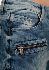 Jeans in denim blu con una texture sbiadita, dotati di una tasca con zip in metallo, e un design classico con cuciture tradizionali.