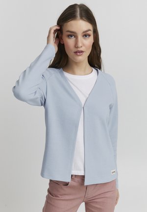 Cardigan côtelé bleu clair avec un col en V et une fermeture éclair à l'avant, porté par-dessus un t-shirt blanc. Il dispose d'une petite étiquette logo en bas.