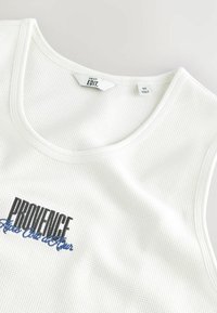 Fehér ujjatlan top, bordás anyagból, amelyen fekete és kék "PROVENCE" szöveg található az elülső részen. A címke közepes méretet jelöl.
