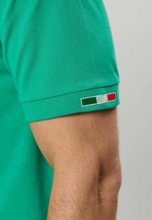 Mouw van een groene T-shirt met korte mouwen met een klein Italiaans vlaggenpatchje vlakbij de zoom, gedragen op de arm van een persoon tegen een neutrale achtergrond.