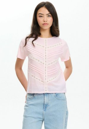 Joven mujer con cabello largo y oscuro que lleva una blusa de manga corta color rosa claro con detalles de encaje y vaqueros azul claro sobre un fondo blanco.
