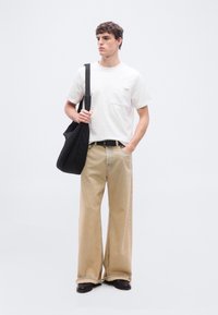 Νέος άνδρας που φοράει λευκό μπλουζάκι και φαρδιά μπεζ τζιν, κρατώντας μια μαύρη τσάντα ώμου, στέκεται μπροστά σε ένα απλό ανοιχτό φόντο.