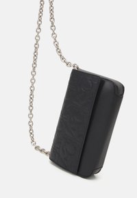 Borsetta a tracolla in pelle nera con lettere in rilievo sulla patta e una tracolla in catena argentata su sfondo bianco.