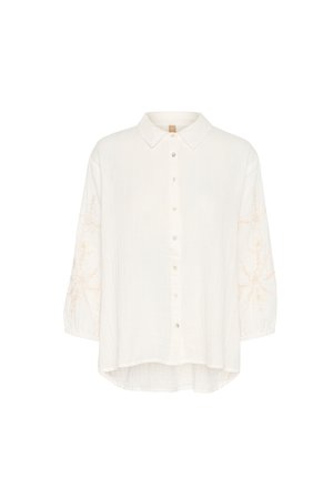 Balts krekls ar apkakli, ar garām piedurknēm un gaiši bežiem izšuvumiem uz piedurknēm. Mīksta, teksturēta auduma.