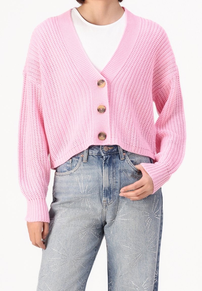Rosa gestrickte Strickjacke mit V-Ausschnitt, die drei große Knöpfe auf der Vorderseite hat. Über einem weißen T-Shirt getragen und mit hellblauen Jeans kombiniert.