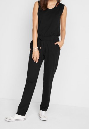 Femme portant une combinaison noire sans manches avec des boutons marron sur l'épaule, des poches latérales, associée à des baskets blanches et des bracelets.