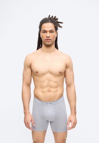 Άνδρας γυμνασμένος με dreadlocks στέκεται στραμμένος προς τα εμπρός, φορώντας ανοιχτό γκρι μποξέρ Calvin Klein, πάνω σε απλό λευκό φόντο.