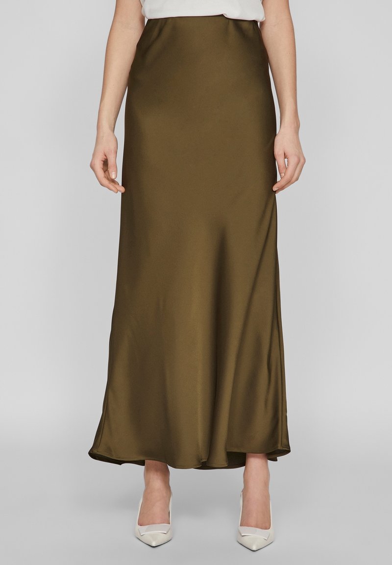 Femme portant une jupe maxi en satin vert olive associée à des talons blancs pointus, debout devant un fond gris uni.