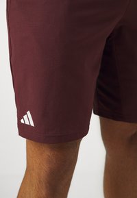 Shorts de sport bordeaux en tissu texturé. Présentent un logo blanc sur l'ourlet inférieur. Coupe au-dessus du genou.