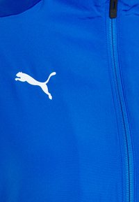 Blue athletic jacket with a smooth texture, featuring a white Puma logo. Includes a zip closure and stitched seams along the front.