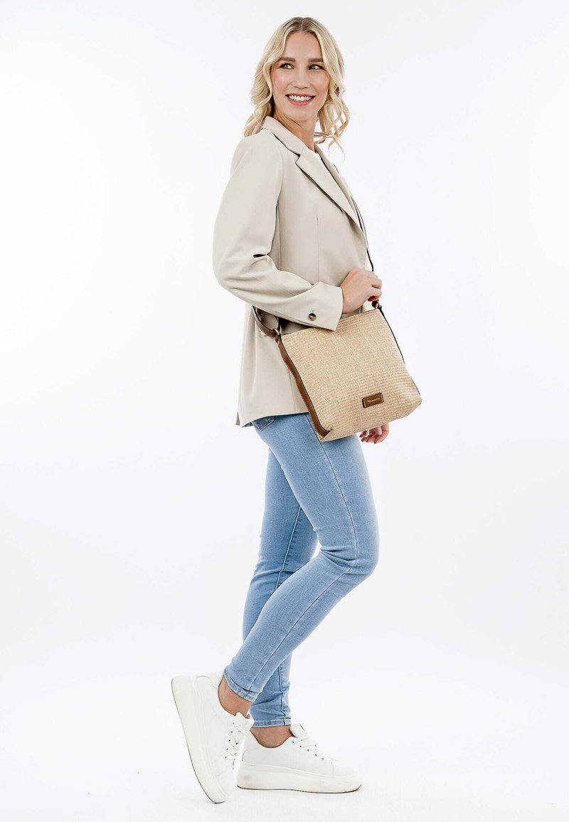 Borsa a spalla intrecciata beige con rifiniture in pelle marrone, forma rettangolare e tracolla regolabile. Il modello indossa un blazer crema e jeans blu.