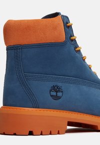 Stivale blu in suede con colletto in suede arancione, dotato di lacci gialli e suola in gomma con un logo ad albero impresso sul lato.