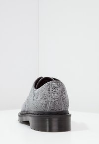 Chaussure en tissu texturé gris avec un motif paisley et une épaisse semelle noire à rainures, photographiée de l'arrière sur une surface et un fond blancs.