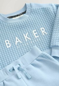 Lyseblå sweatshirt og sweatpants sæt. Tekstureret top har "BAKER" i hvid, med knapdetaljer og snor i taljen. Blødt stof.